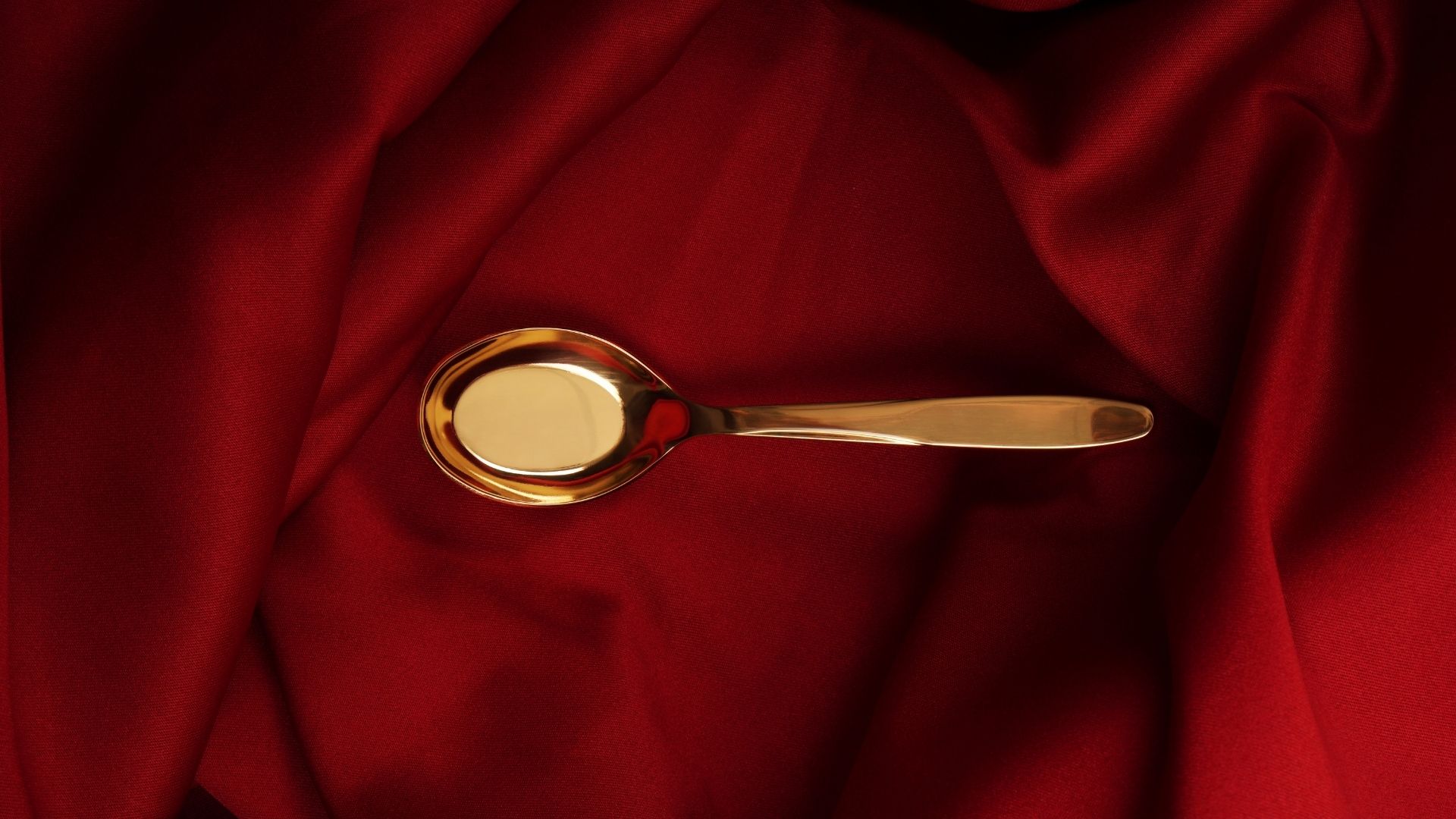 The photo showcases a sleek Ladata gold-plated Taiwanese spoon resting on a rich red fabric background, highlighting its shiny, reflective surface and elegant design. The deep red texture of the fabric contrasts beautifully with the spoon's metallic sheen, emphasizing its luxurious appeal.