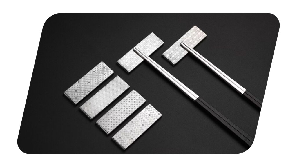 The image displays six stainless steel chopstick rests with intricate geometric patterns, arranged on a dark background. Two of the rests are paired with sleek chopsticks, one with a metallic finish and the other with a black handle, creating a modern and elegant presentation.