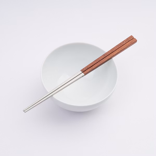 The image shows a pair of chopsticks with a metallic section and a wooden handle in a warm brown tone, resting diagonally across a simple white bowl. The setup is placed against a plain white background, emphasizing the clean and minimalist design of the utensils.