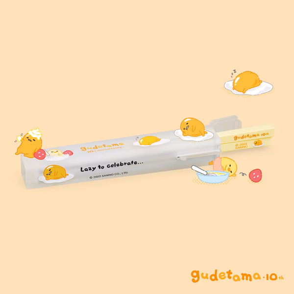 The image features a set of Gudetama-themed portable BaoQuai chopsticks housed in a clear case with a playful design, showcasing the lazy egg character in various poses along with the text "Lazy to celebrate..." and "Gudetama 10th" The case is decorated with cute illustrations of Gudetama, some with food items, set against a soft beige background, highlighting the fun and whimsical theme.